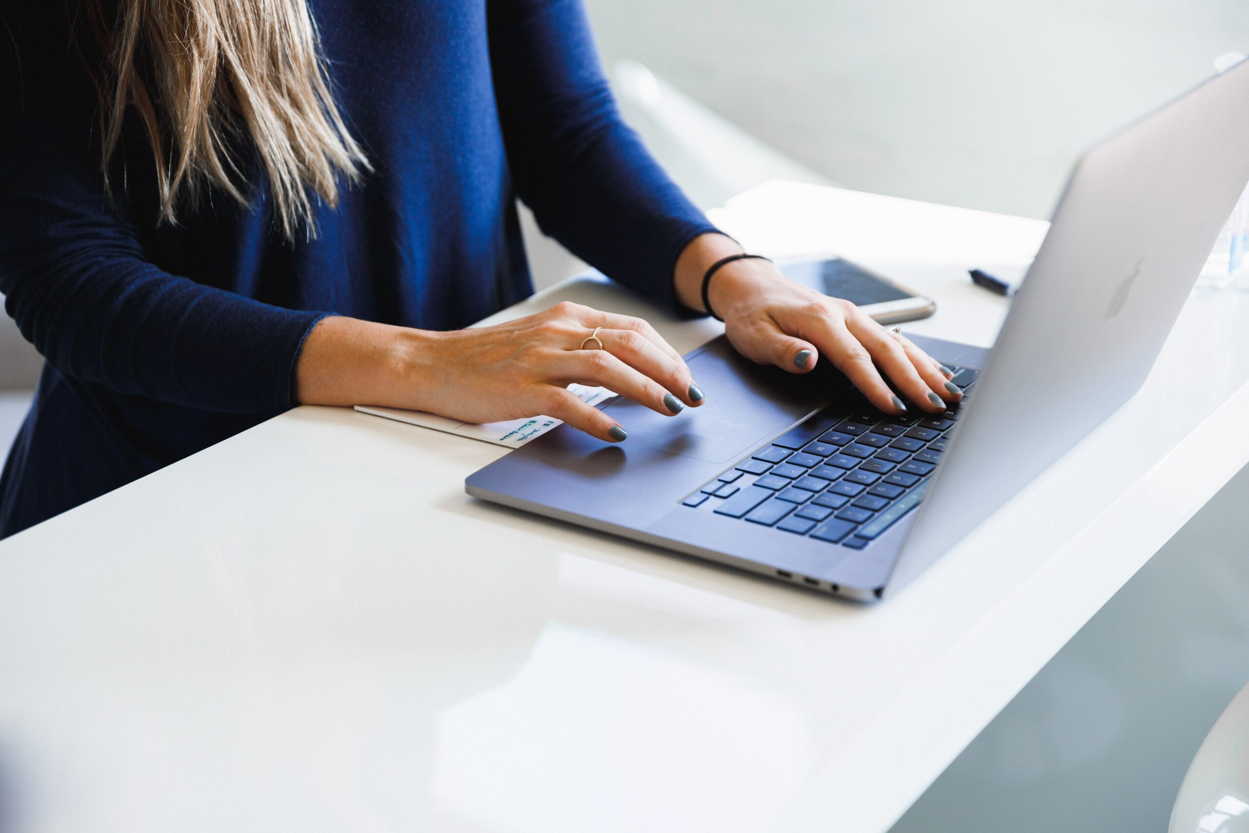 Woman-in-a-blue-top-sitting-at-a-white-desk-working-on-a-laptop-scaled